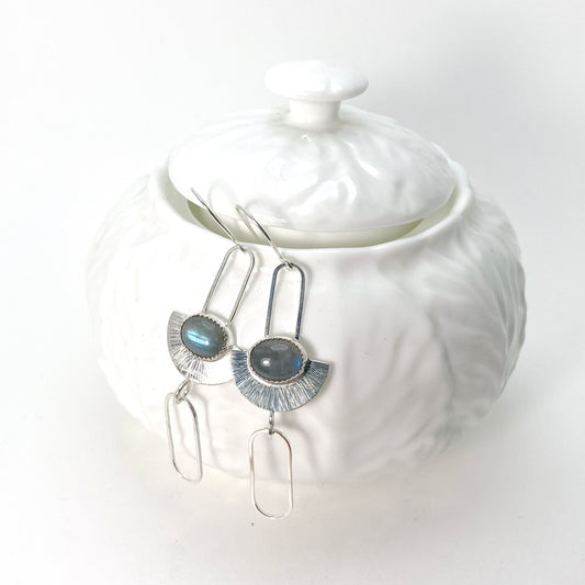 Sterling silver earrings in an art-deco geometric style. An oval labradorite stone lays horizontally across a half-circle with radial lines. The half-circle is suspended from a hook by a skinny, elongated square-wire half chain link. Below the half-circle, another elongated chainlink swing freely from a jump ring.