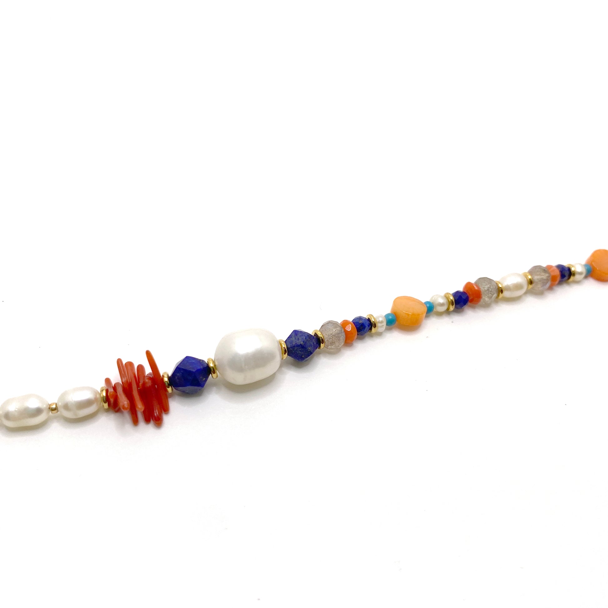 Asymmetrical peal and ocean-inspired gemstone necklace. Just over half of the necklace is made from small oblong freshwater pearls interspersed with gold seed beads. The rest is a pattern of vintage coral, lapis lazuli, pearl, labradorite, and gold and turquoise coloured spacer beads.